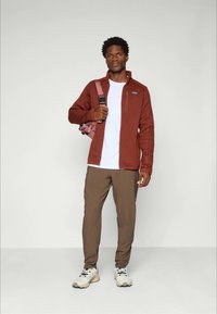 Jachetă maro din fleece, tricou alb, pantaloni maronii și adidași de culoare deschisă. Modelul ține un rucsac roz, stând în fața unui fundal deschis.