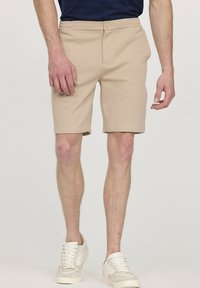 Shorts beiges en tissu doux avec une taille élastique, fermeture à bouton sur le devant et poches latérales, associés à des baskets blanches.
