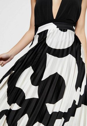 Jupe plissée noire et blanche avec des motifs abstraits audacieux portée avec un haut noir à col en V profond, laissant voir une partie des bras et du torse.