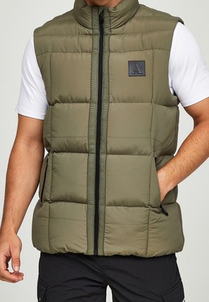 Homme portant un gilet sans manches matelassé vert olive sur une chemise blanche à manches courtes avec un pantalon cargo noir, une main dans la poche du gilet.