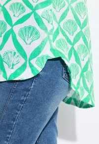 Grünes und weißes gemustertes Hemd mit Muschelmotiven, aus leichtem Stoff gefertigt, kombiniert mit blauen Jeans. Der Saum hat eine leichte Rundung.