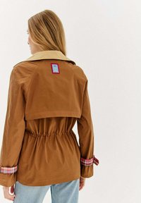 Hnedá bunda s béžovým golierom, elastickým pásom a károvanými manžetami. Na zadnej strane má modrý a červený nášivku. Hladká látka a štruktúrovaný dizajn.