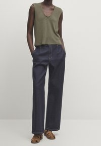 Olive green knitted tank top with a scoop neckline, paired with loose dark blue denim trousers and brown strappy sandals.