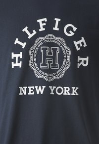 Námorně modré bavlněné tričko s velkým bílým nápisem "HILFIGER" a "NEW YORK", s kruhovým znakem uprostřed, který zdůrazňuje písmeno "H."