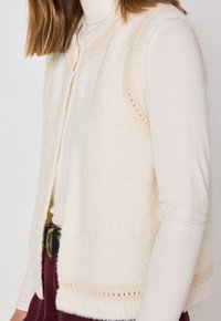 Gilet texturé de couleur crème avec une ouverture sur le devant, tissu doux et détails en dentelle. Porté sur un haut à manches longues de couleur claire.