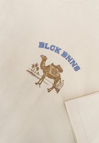Beige katoenen t-shirt met een graphic van een kameel met palmbomen en piramides, tekst "BLCK BNNS" in het blauw boven het ontwerp.