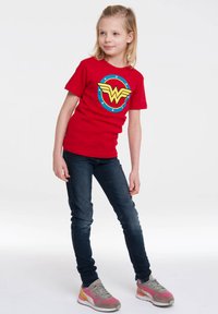 Red cotton t-shirt featuring a yellow and blue Wonder Woman logo, paired with dark blue jeans and multicolored sneakers.