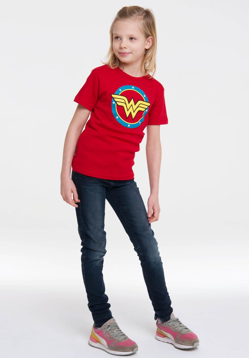 Red cotton t-shirt featuring a yellow and blue Wonder Woman logo, paired with dark blue jeans and multicolored sneakers.