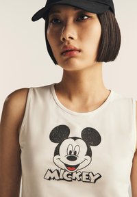 Giovane donna con capelli corti e scuri che indossa un cappellino nero e un top senza maniche bianco con una grafica di Topolino.