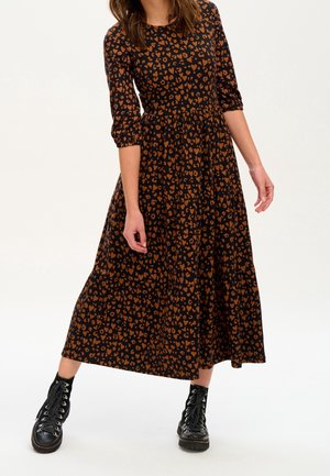 Frau trägt ein schwarzes Midikleid mit braunem Herzenmuster und dreiviertel Ärmeln, kombiniert mit schwarzen Schnürstiefeln, steht vor einem schlichten Hintergrund.