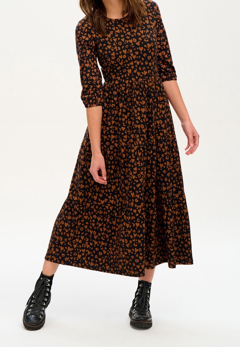 Frau trägt ein schwarzes Midikleid mit braunem Herzenmuster und dreiviertel Ärmeln, kombiniert mit schwarzen Schnürstiefeln, steht vor einem schlichten Hintergrund.