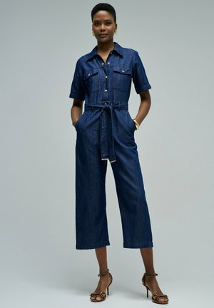 Mujer de pie con las manos en los bolsillos, vistiendo un mono de mezclilla azul oscuro y sandalias marrones de tacón alto, contra un fondo gris claro.