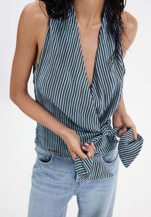 Femme portant un haut cache-cœur sans manches à rayures bleues et noires noué à la taille, associé à un jean bleu clair, sur fond blanc.