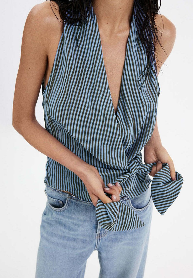 Femme portant un haut cache-cœur sans manches à rayures bleues et noires noué à la taille, associé à un jean bleu clair, sur fond blanc.