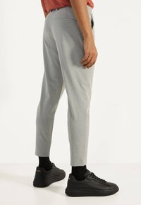 Man som bär ljusgrå slim-fit byxor, svarta strumpor och svarta sneakers, stående mot en vit bakgrund.