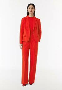 Red tailored suit with a fitted blazer and straight-leg trousers, made of smooth fabric. High-heeled red shoes add a bold accent.