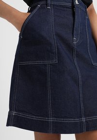 Falda de mezclilla oscura con grandes bolsillos frontales, costuras blancas contrastantes, una silueta recta y un dobladillo que cae a mitad del muslo.