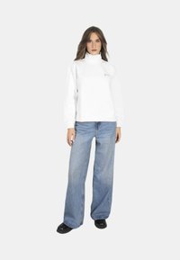 Felpa bianca a collo alto con logo, abbinata a jeans blu a sigaretta e scarpe nere. Tessuto morbido, stile casual.