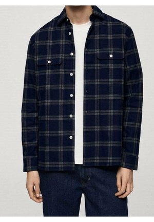 Homme portant une chemise à carreaux bleu marine et grise boutonnée sur un t-shirt blanc avec un jean bleu foncé, debout, les mains détendues.