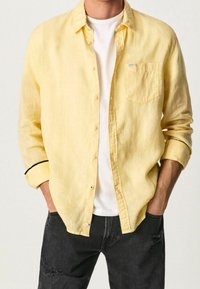 Camisa de lino amarilla con botones y un bolsillo en el pecho, con detalles negros en las mangas, llevada sobre una camiseta blanca y combinada con unos jeans negros desgastados.