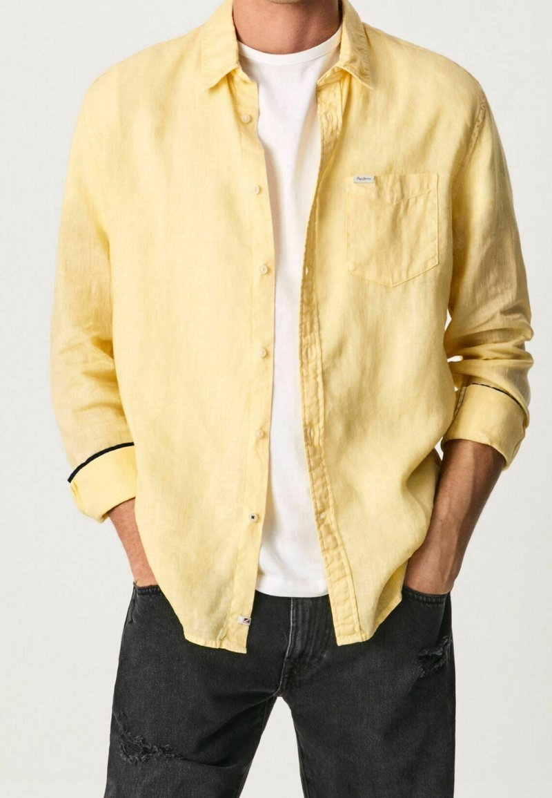 Camisa de lino amarilla con botones y un bolsillo en el pecho, con detalles negros en las mangas, llevada sobre una camiseta blanca y combinada con unos jeans negros desgastados.