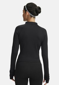 Vrouw met donker haar in een bun, gekleed in een zwart langslijf-top en zwarte leggings, met haar rug naar de camera gericht tegen een effen lichte achtergrond.