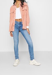 Camisa de terciopelo de cordón color coral con dos bolsillos en el pecho, combinada con jeans ajustados de color azul claro y zapatillas blancas. Diseño simple y casual.