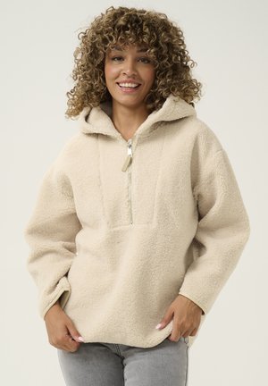 Beige fleece pullover met een halve rits, capuchon en textuurstof. Heeft een kangoeroezak en verlaagde schouders voor een ontspannen pasvorm.