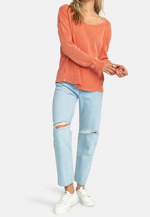 Top coral de manga larga con costillas y corte oversize, combinado con jeans azules claros rasgados y zapatillas blancas. Detalles destacados de estilo relajado.