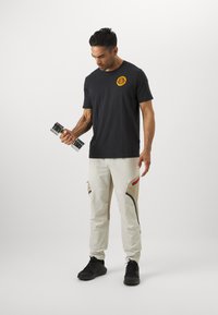 T-shirt noire avec logo orange, pantalon clair avec accents rouges et poches, tenant un haltère en argent, baskets noires.