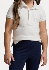 Biała koszulka polo z krótkim rękawem, teksturowana tkanina, trzy guziki przy dekolcie, małe różowe logo, zestawiona z ciemnoniebieskimi legginsami.