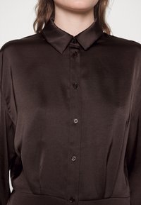 Chemise en satin marron avec un col classique, une fermeture à boutons avec des boutons foncés, et une texture lisse. Aucune motif ou accent notable.