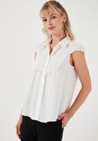 Bílá blůza s krátkými nafouklými rukávy, zapínáním na knoflíky a složitým krajkovým zdobením na přední straně. Lehký materiál s volným střihem.