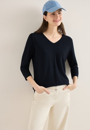Femme souriante, portant une casquette bleu clair, un chemisier noir à col en V à manches longues, et un pantalon beige, debout avec les mains dans les poches contre un mur uni.