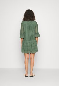 Green lace dress with three-quarter sleeves, detailed embroidery, a flared hem, and a relaxed fit. Worn over simple sandals.