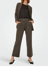 Kvinna klädd i en mörk olivgrön skräddarsydd blazer och vida byxor med en svart blus och svarta spetsiga klackskor, stående med händerna i fickorna.