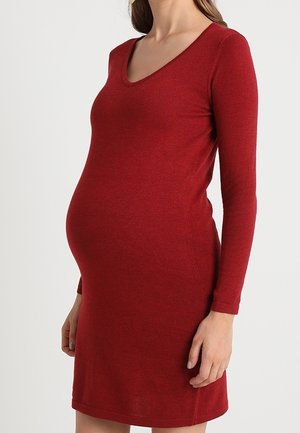 Femme enceinte portant une robe rouge ajustée à manches longues avec un décolleté arrondi, debout devant un fond clair uni.