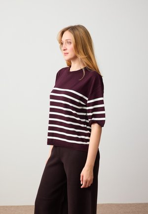 Femme aux longs cheveux blonds portant un haut à manches courtes rayé violet et blanc, ainsi qu'un pantalon noir, debout de profil devant un fond uni.