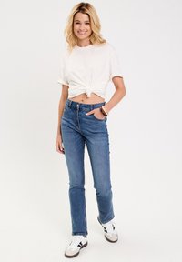 Hvid knyttet t-shirt med op-rullede ærmer, parret med mellemhøje blå jeans. Sneakers har en hvid base med sorte og grå detaljer.