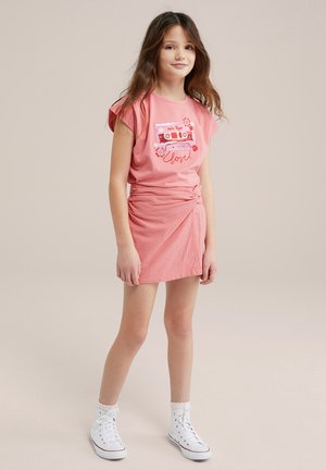Rosa T-Shirt mit einem Grafikdesign von einem Kassette und floralen Akzenten. Wickelrock aus strukturiertem Pink. Weiße High-Top-Sneaker mit gewellten Socken.