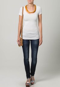 Bijela prilagođena majica s zlatnim izrezom, uparen s tamnoplavim trapericama i smeškim preko ramena. Model nosi sivo visoke cipele.