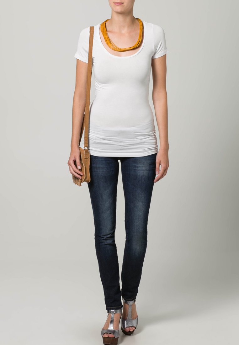 Bijela prilagođena majica s zlatnim izrezom, uparen s tamnoplavim trapericama i smeškim preko ramena. Model nosi sivo visoke cipele.