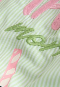 Zelený vyšívaný nápis "mom" na světle zelené a bílé pruhované látce s růžovými a bílými dekorativními prvky.