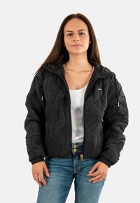 Zwarte gewatteerde jas met gegraveerde patronen, ritssluiting aan de voorkant, verstelbare capuchon en elastische manchetten. Gdragen over een witte top en denim broek.