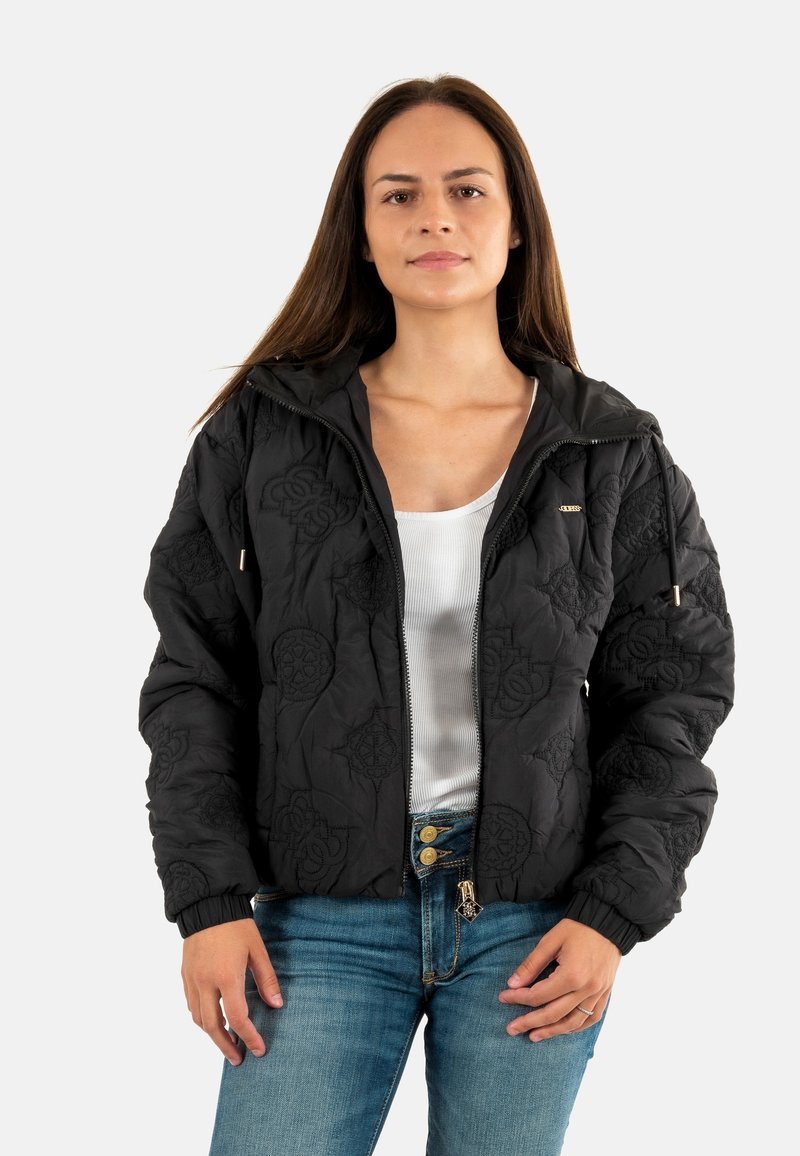 Zwarte gewatteerde jas met gegraveerde patronen, ritssluiting aan de voorkant, verstelbare capuchon en elastische manchetten. Gdragen over een witte top en denim broek.