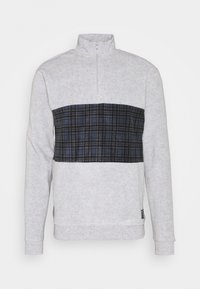 Grå fleece-tröja med dragkedja i kragen, med ett svart och blått rutmönster över bröstet. Mjuk textur och ribbade muddärmar.