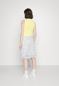 Žlutý svrchní top bez rukávů s pleteným vzorem ve tvaru kabelky, kombinovaný s bílou sukni s květinovým vzorem a volány, obutý s bílými teniskami a kotníkovými ponožkami, pohled zezadu.