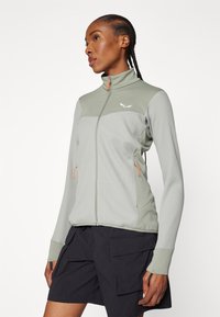 Giacca zip-up verde chiaro con tessuto texturizzato, pannelli grigi a contrasto, zip arancioni e un design aderente. Abbinata a pantaloni cargo neri.