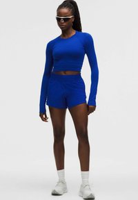 Zilā ilgās piedurknes krekliņa un šortu komplekts ar pieguļošu dizainu un gludu tekstūru. Aksesuāri ietver pelēkas sporta apavus un baltas zeķes līdz potīti.