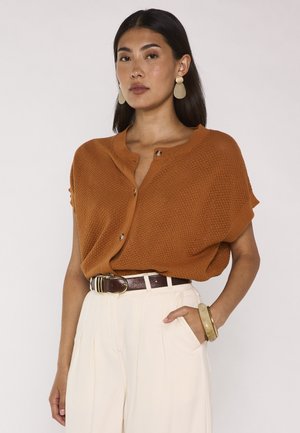 Frau trägt ein rostfarbenes kurzärmeliges Strickoberteil mit Knöpfen, cremefarbene Hose mit hoher Taille, braunen Gürtel, beige Ohrringe und Armreifen an einem Handgelenk.